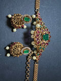 A piece from Saanskrityam Handicraft Store's collection, the Jewellery/Indian cosmetic jewellery with stones and metal, featuring a gold chain entwined with vibrant green gemstones reminiscent of traditional Indian style, is beautifully draped over a textured wooden surface. The muted dark background enhances the contrast and richness of this exquisite piece.