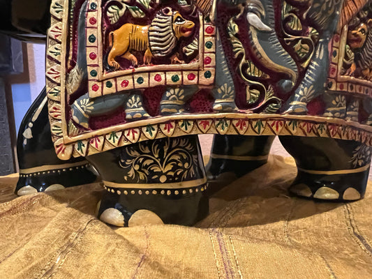 A beautifully crafted piece from the Saanskrityam Handicraft Store, this wood carved elephant sculpture features an ornate red and gold design with intricate patterns and a raised trunk. It rests on a yellow surface, showcasing its Indian-inspired style with colorful floral motifs, stripes, and a round ornament on its back that enhances the traditional elegance typical of Indian elephant carvings.