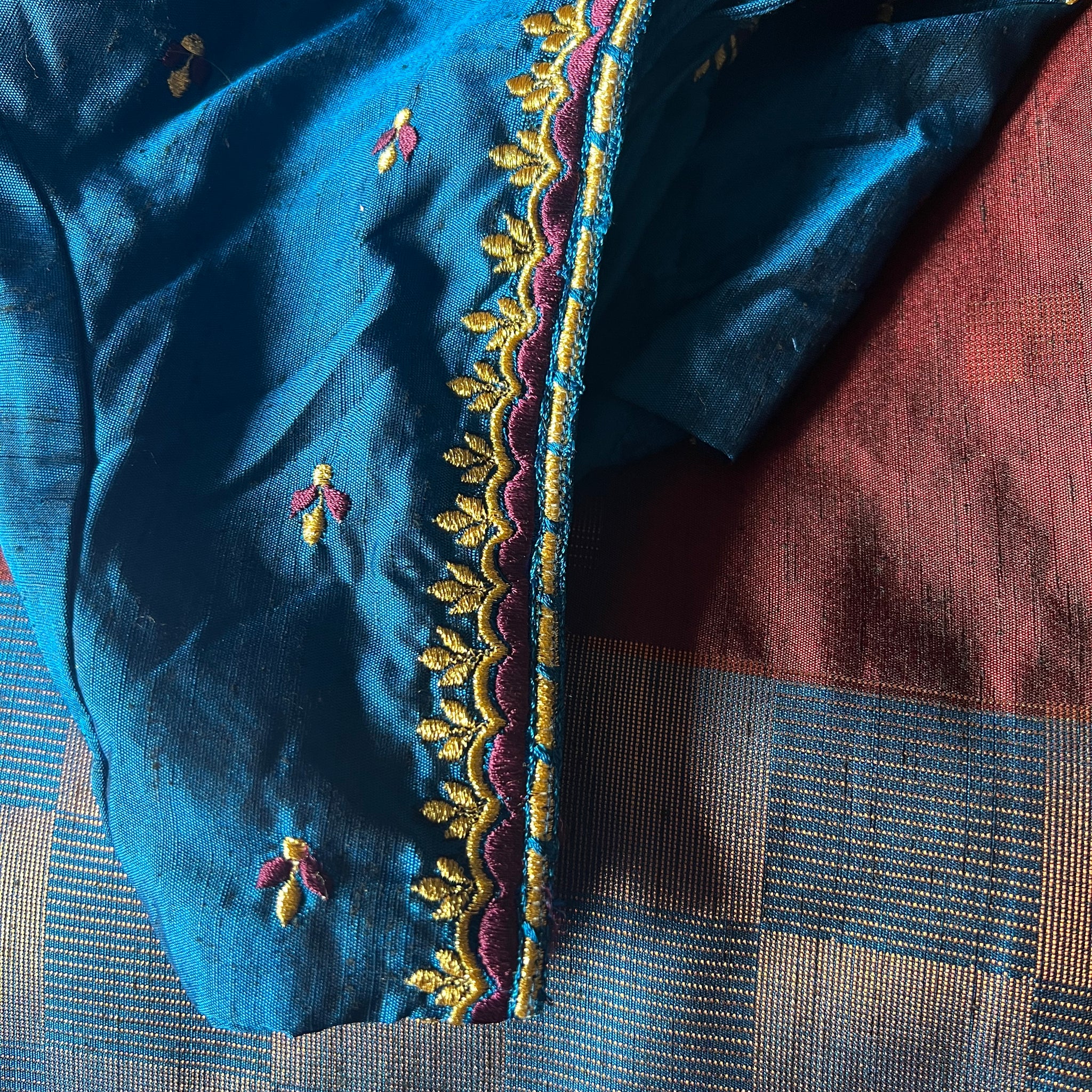 A vibrant blue semi-silk saree from the Saanskrityam Handicraft Store, featuring intricate gold and pink embroidery with floral motifs and a scalloped border, is beautifully draped over a contrasting red and blue checked surface. It pairs perfectly with trendy blouses, and comes with a matching embroidered blouse in size large.