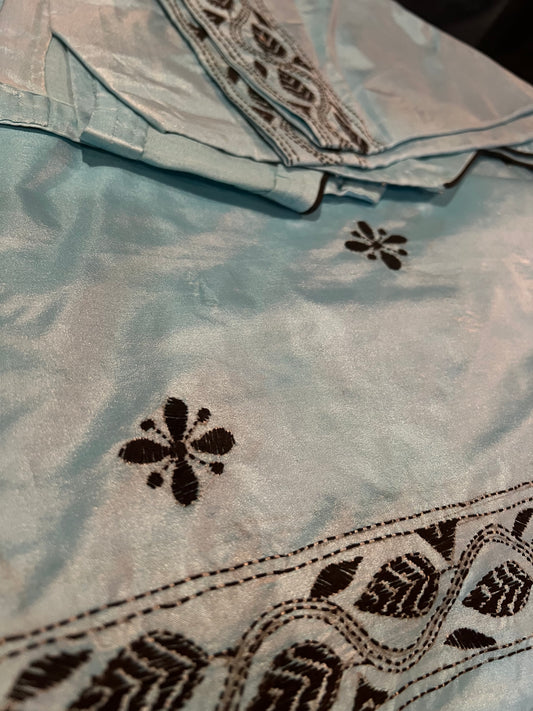 Detailed view of the Kantha thread work Bangla silk saree from Saanskrityam Handicraft Store, featuring intricate black threadwork that forms elegant floral and vine patterns on a light blue background. The shiny texture enhances its appeal, with a folded section at the top. This fabric includes a ready-to-wear blouse, making it an excellent choice for sophisticated attire.