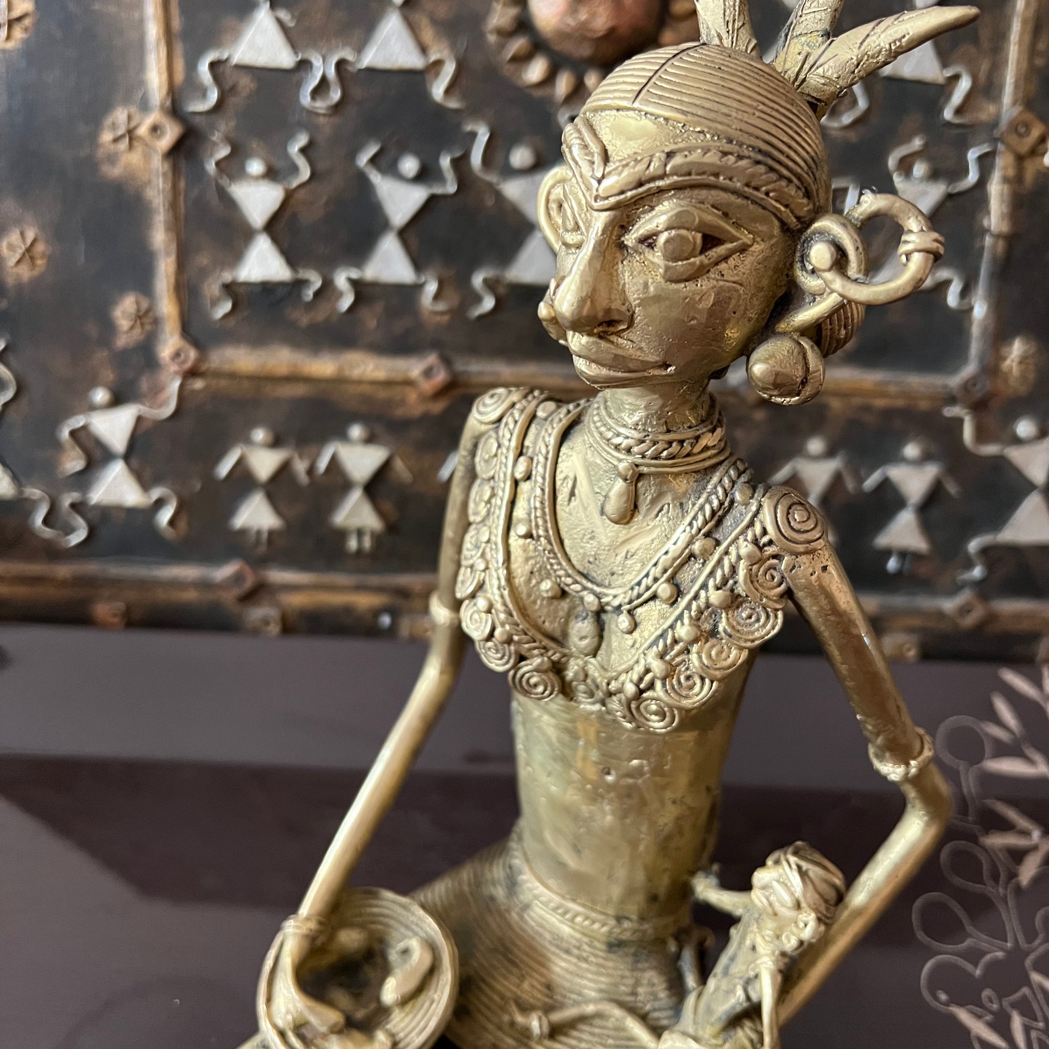 A bronze Chhattisgarh art sculpture from the Saanskrityam Handicraft Store presents a seated tribal woman, adorned with intricate jewelry and a headdress, holding a small object. The background includes a decorative metal panel embellished with geometric patterns and a sun motif.