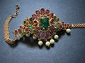 A piece from Saanskrityam Handicraft Store's collection, the Jewellery/Indian cosmetic jewellery with stones and metal, featuring a gold chain entwined with vibrant green gemstones reminiscent of traditional Indian style, is beautifully draped over a textured wooden surface. The muted dark background enhances the contrast and richness of this exquisite piece.