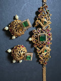 A piece from Saanskrityam Handicraft Store's collection, the Jewellery/Indian cosmetic jewellery with stones and metal, featuring a gold chain entwined with vibrant green gemstones reminiscent of traditional Indian style, is beautifully draped over a textured wooden surface. The muted dark background enhances the contrast and richness of this exquisite piece.