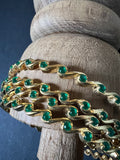 A piece from Saanskrityam Handicraft Store's collection, the Jewellery/Indian cosmetic jewellery with stones and metal, featuring a gold chain entwined with vibrant green gemstones reminiscent of traditional Indian style, is beautifully draped over a textured wooden surface. The muted dark background enhances the contrast and richness of this exquisite piece.