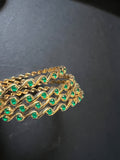 A piece from Saanskrityam Handicraft Store's collection, the Jewellery/Indian cosmetic jewellery with stones and metal, featuring a gold chain entwined with vibrant green gemstones reminiscent of traditional Indian style, is beautifully draped over a textured wooden surface. The muted dark background enhances the contrast and richness of this exquisite piece.