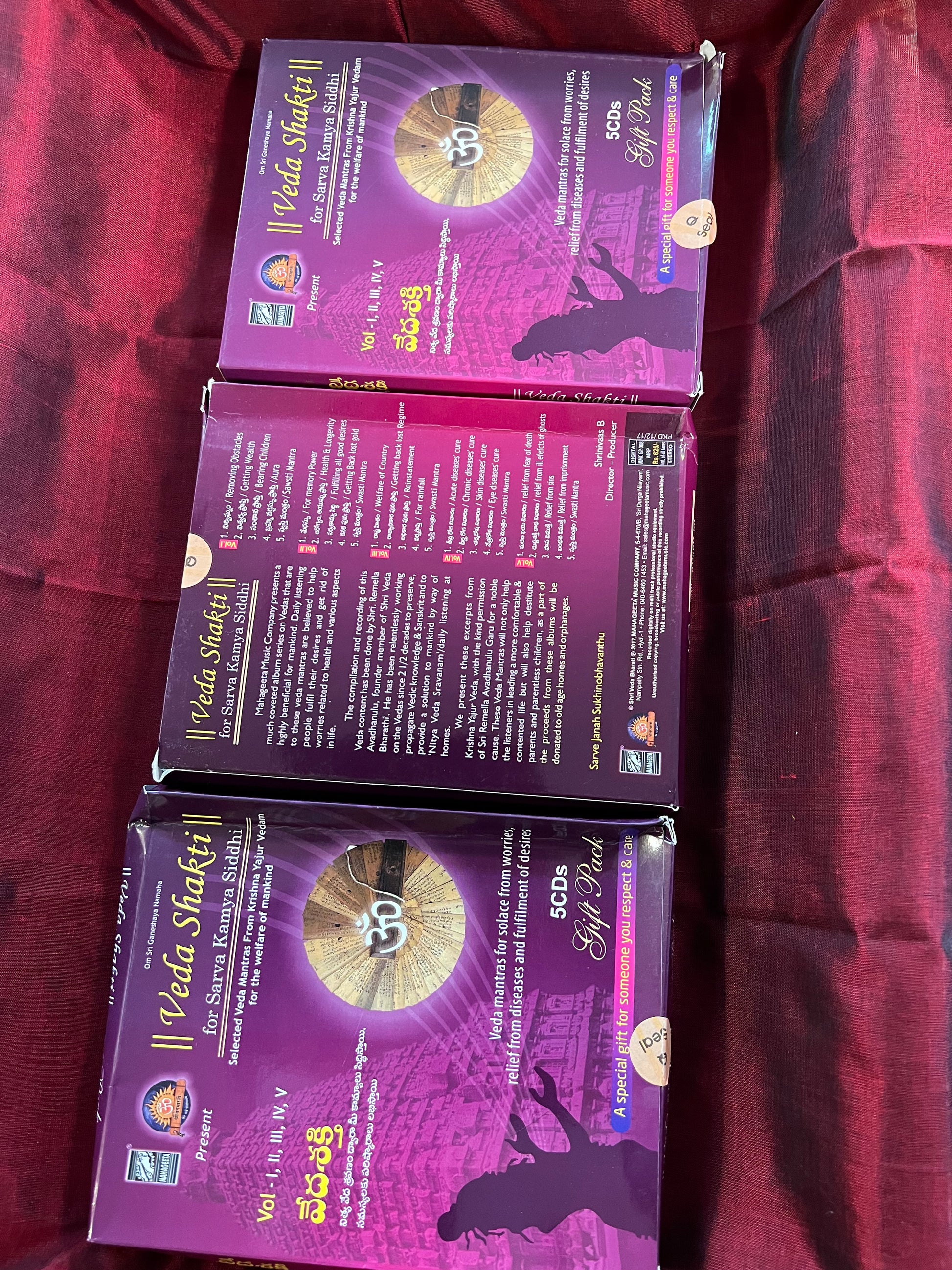 Three packages of the Indian Music Collection incense sticks from Saanskrityam Handicraft Store are arranged on a red fabric. The purple boxes with gold text, featuring an OM symbol and various logos, evoke rich fragrances akin to the soothing notes of Carnatic music.